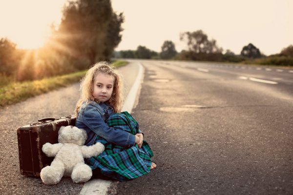 child sitting on side of road with suitcase and teddy bear